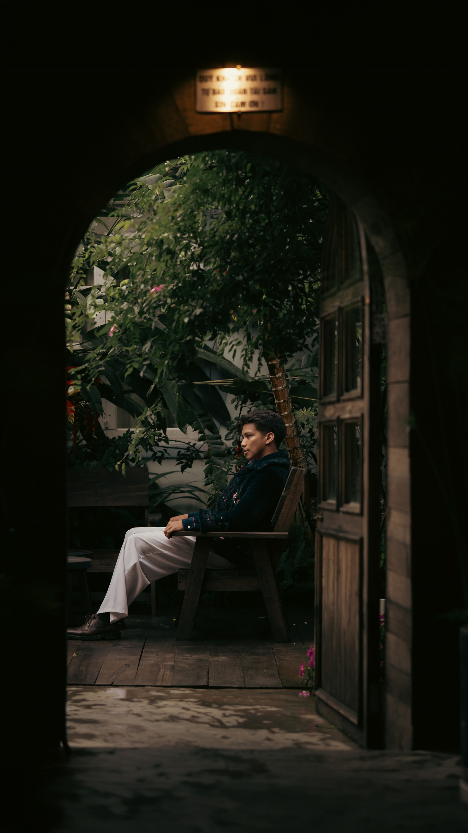 Lou Villaluna seated in an arched wooden doorway, a garden behind him